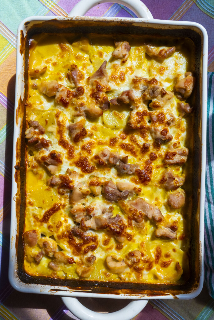 A baked casserole dish filled with golden-brown, cheesy, and bubbly cooked chicken pieces on top of layers of potatoes and creamy sauce, resting on a colorful, striped tablecloth.