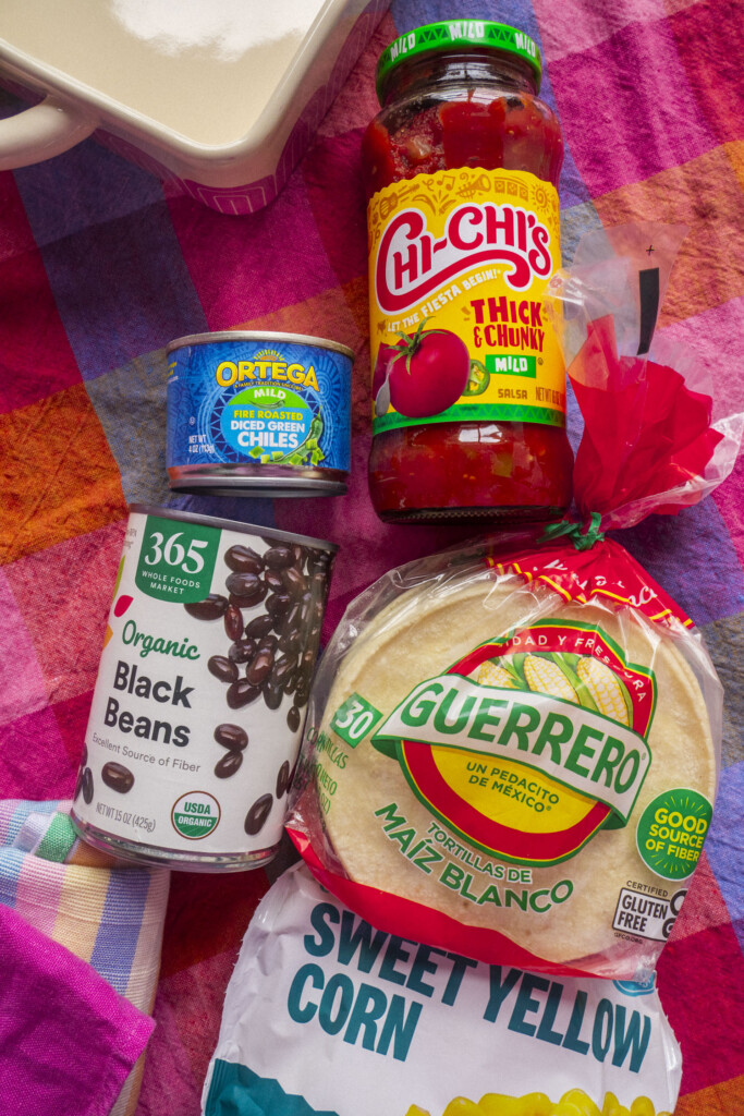A can of black beans, a can of diced green chiles, a jar of chi chi's salsa, a package of corn tortillas, and a bag of sweet yellow corn are arranged on a colorful checkered tablecloth.