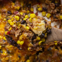 A close-up of a wooden spoon holding a serving of casserole with melted cheese, black beans, corn, ground meat, and diced tomatoes. The casserole appears cheesy and baked, with visible golden brown edges.