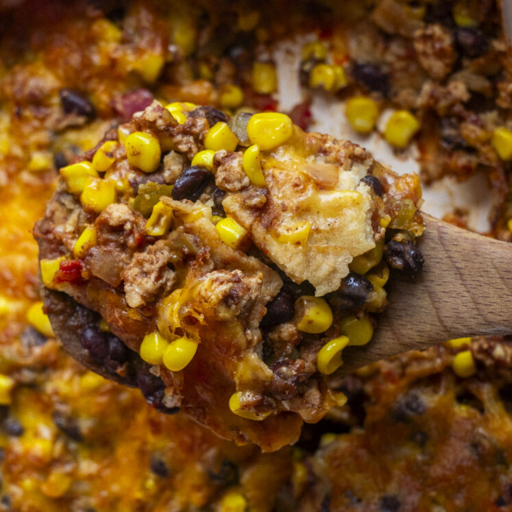 A close-up of a wooden spoon holding a serving of casserole with melted cheese, black beans, corn, ground meat, and diced tomatoes. The casserole appears cheesy and baked, with visible golden brown edges.