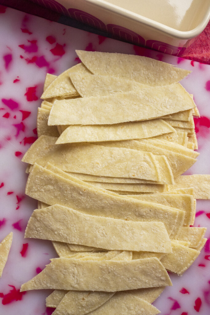 Several uncooked corn tortillas cut into strips are arranged on a pink and white marbled cutting board, with a beige baking dish partially visible in the background.