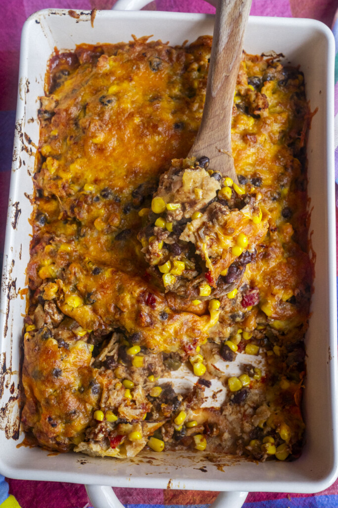 A rectangular baking dish filled with a cheesy, baked casserole containing corn, beans, ground meat, and melted cheese, with a wooden spoon scooping out a portion.
