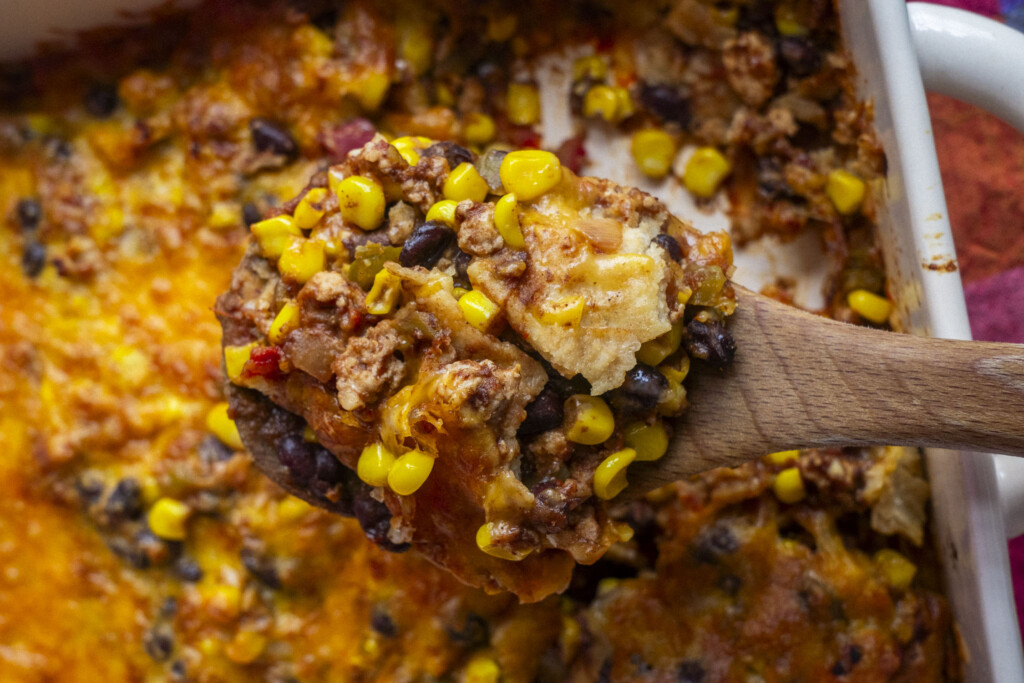 A wooden spoon holds a serving of cheesy casserole with corn, black beans, ground meat, and melted cheese above a baking dish filled with the same colorful mixture.