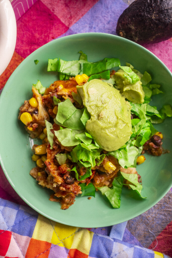 A green bowl filled with a colorful mix of beans, corn, lettuce, and a generous scoop of guacamole, sitting on a vibrant, multicolored cloth with an avocado nearby.