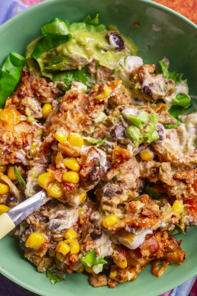 A close-up of a bowl filled with a colorful mixture of corn, black beans, melted cheese, guacamole, lettuce, saucy sour cream and ground meat, with a fork resting inside.
