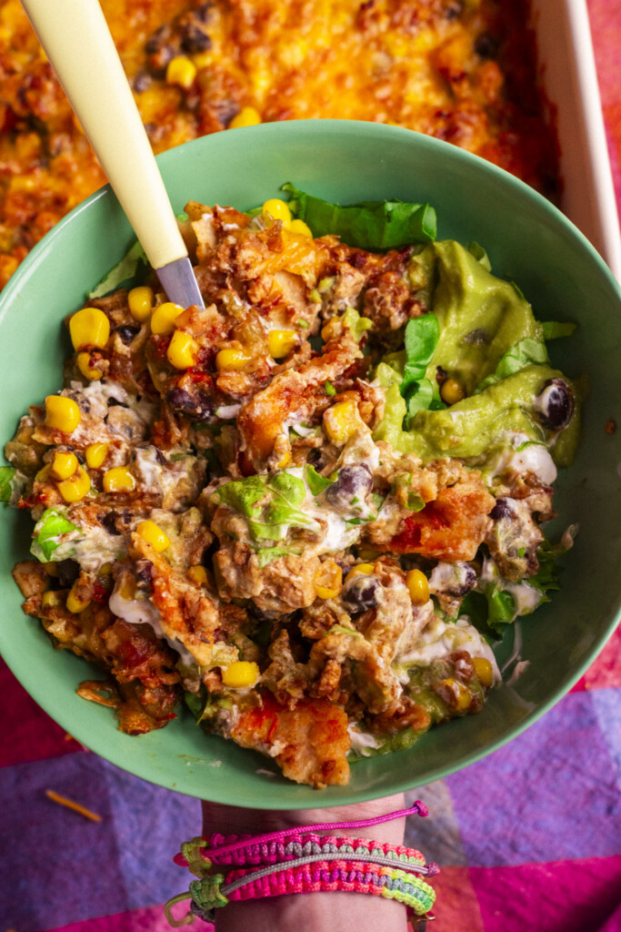 A hand holding a green bowl filled with a cheesy Tex-Mex casserole featuring ground chicken, corn, black beans, lettuce, and guacamole, with a fork in the bowl. The person is wearing colorful woven bracelets.