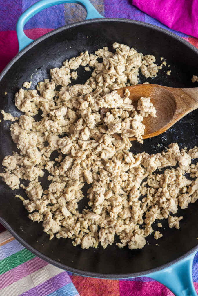 Cooked ground chicken in a black skillet, being stirred with a wooden spoon. The skillet is on a colorful surface with striped and pink towels nearby.