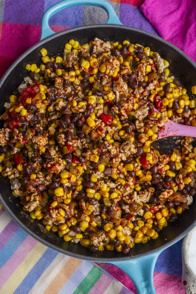 A skillet filled with a colorful mix of corn, black beans, ground meat, and red bell peppers, with a pink spoon on the side. The skillet is on a striped tablecloth with a bright pink napkin.