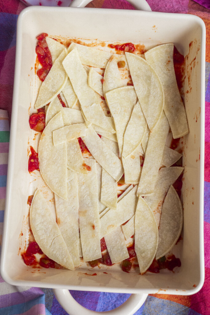 A baking dish with a layer of chopped corn tortillas over a tomato-based sauce, prepared on a colorful tablecloth.
