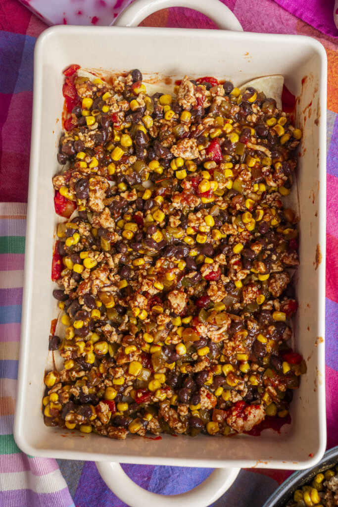 A rectangular baking dish filled with a mixture of ground meat, black beans, corn, tomatoes, and spices, sitting on a colorful tablecloth.
