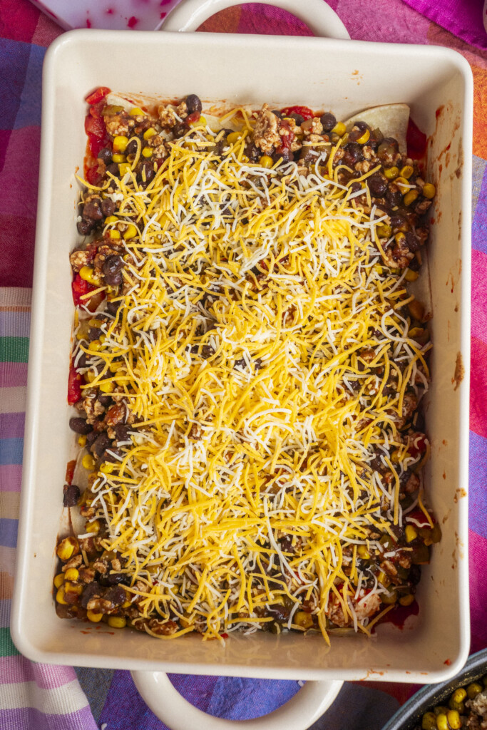 A rectangular baking dish filled with a layered casserole topped with shredded cheese, black beans, corn, ground meat, and diced tomatoes, ready to be baked. The dish is on a colorful tablecloth.