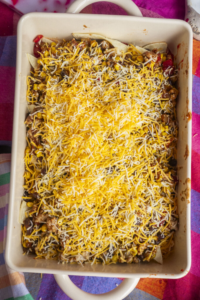 A casserole dish filled with a layered Mexican-style dish topped with shredded yellow and white cheese, ready to be baked. The dish sits on a colorful, checkered tablecloth.