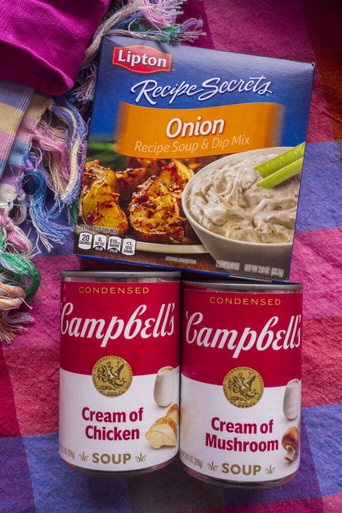 A box of Lipton Recipe Secrets Onion Soup & Dip Mix sits above two cans of Campbells soup—one Cream of Chicken and one Cream of Mushroom—on a colorful plaid fabric background.