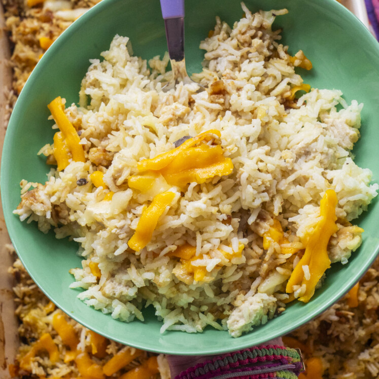 A green bowl filled with rice mixed with shredded chicken and topped with melted cheddar cheese, with a fork resting inside.