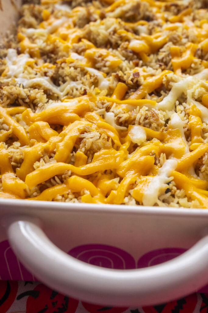 A close-up of a casserole dish filled with cooked rice topped with melted cheddar cheese and bits of crumbly brown soup mix. The dish has a white handle and a pink patterned side.
