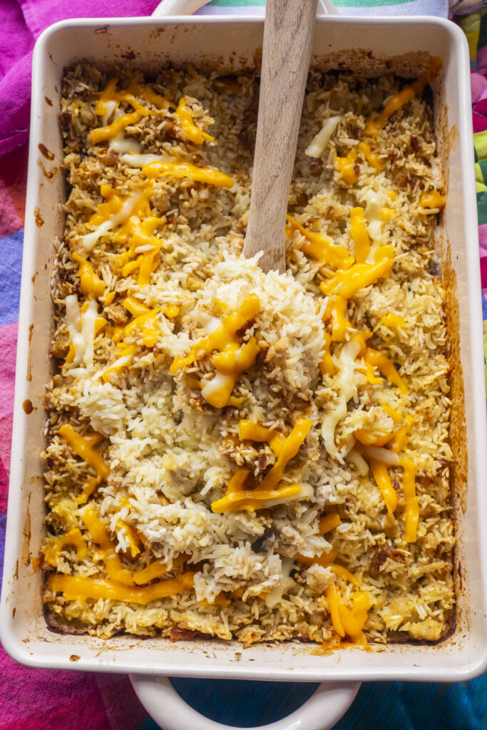 A casserole dish filled with baked rice topped with melted cheddar cheese, with a wooden spoon scooping some out. The edges of the dish are golden and slightly crispy.