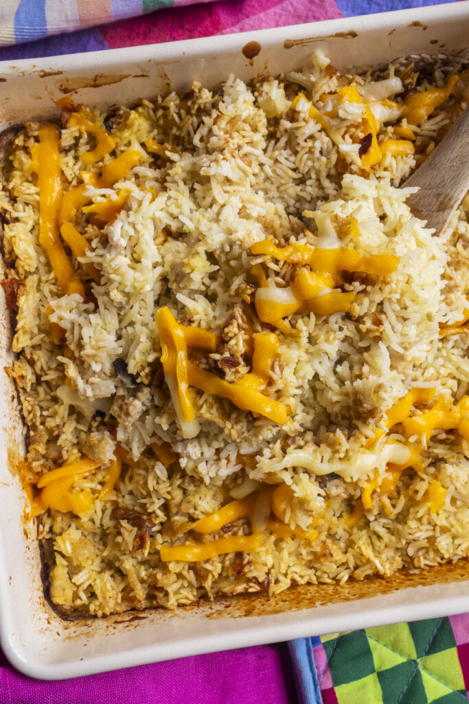 A close-up of a baked casserole dish filled with rice, melted cheddar cheese, chunks of chicken, and a wooden spoon, set on a colorful tablecloth and oven mitt.