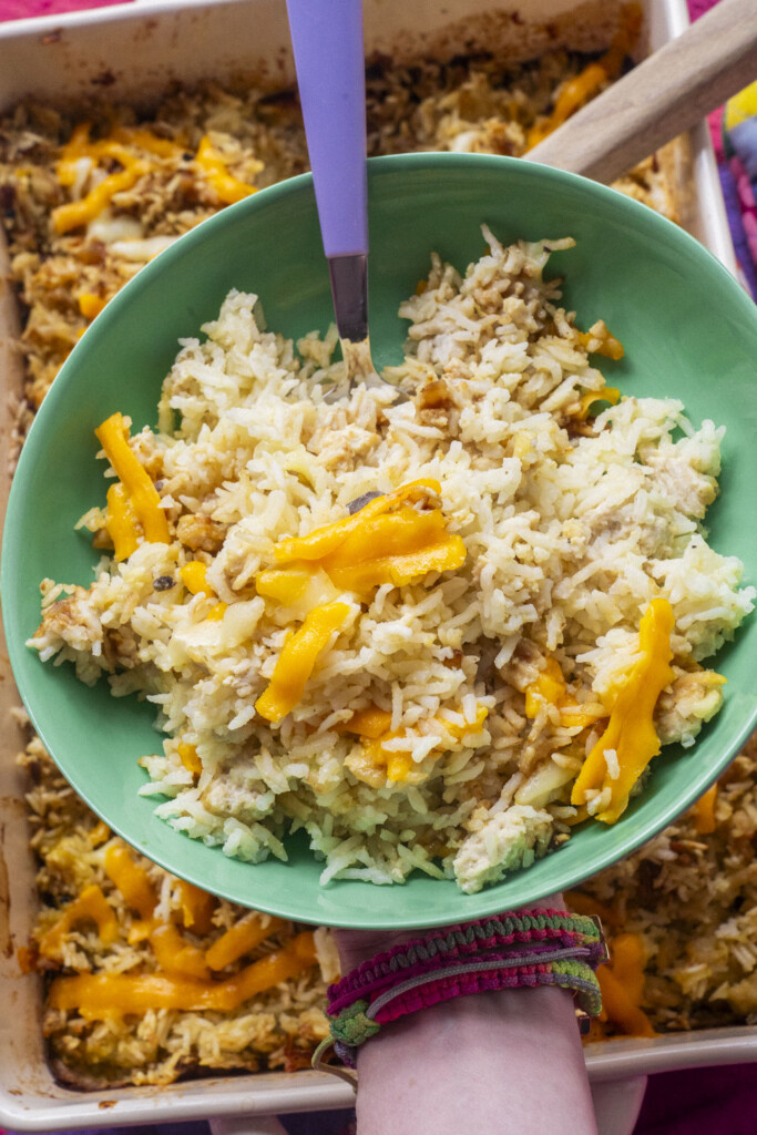 A hand holds a green bowl filled with cooked rice topped with melted cheddar cheese, held above a baking dish of similar rice and cheese casserole. A purple-handled fork rests in the bowl.