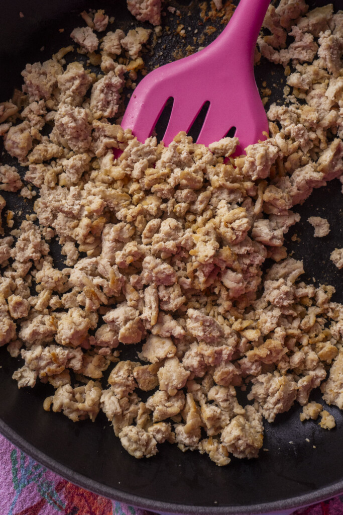 Cooked ground chicken being stirred in a skillet with a bright pink spatula. The meat appears to be well-browned and crumbled, and the pan is placed on a colorful cloth.