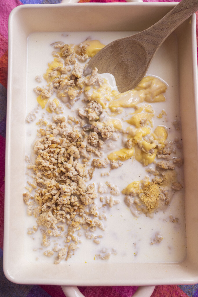 A baking dish containing milk, crumbled chicken, soups, milk and a wooden spoon, set on a colorful cloth. The ingredients appear to be ready for mixing.