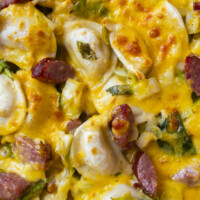 Close-up of a cheesy baked dish featuring golden-brown melted cheese, sliced sausage, pierogi, and pieces of green vegetables, all mixed and covered in a rich, creamy sauce.