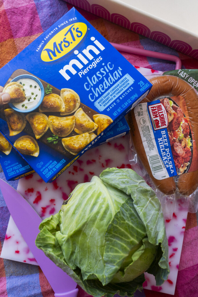 A box of Mrs. T’s mini classic cheddar pierogies, a Kielbasa Polska sausage, a head of cabbage, and a pink knife on a white and red cutting board, all placed on a colorful plaid cloth.