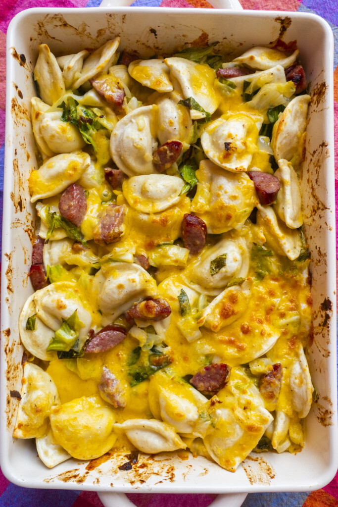 A baked casserole dish filled with pierogi, sliced sausage, melted cheddar cheese, and chopped green onions, with golden brown spots on top and browned edges on the dish.
