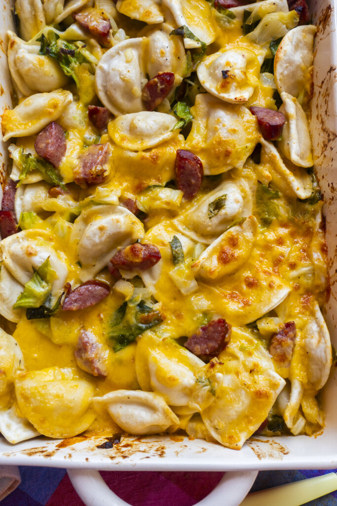 Baked pierogi casserole with melted cheddar cheese, sliced sausage, and green herbs in a white dish. The cheese is golden and bubbly, covering the pierogi and sausage pieces.