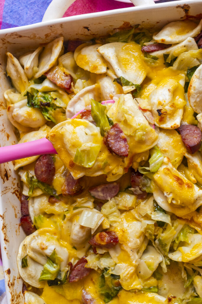 A close-up of a cheesy baked casserole with sliced sausage, cabbage, and pierogi, being scooped from a white baking dish with a pink spatula.