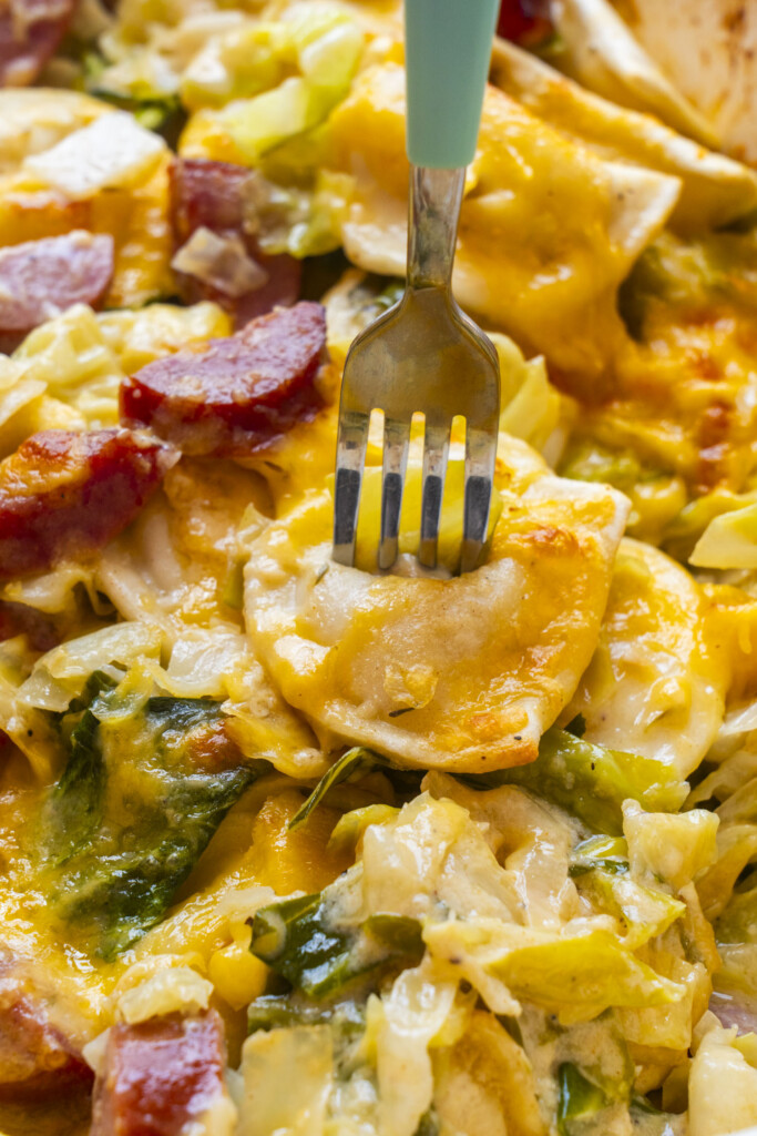 A fork is piercing a cheesy dish with pierogies, sliced sausage, melted cheese, greens, and creamy sauce. The food looks rich, colorful, and comforting.