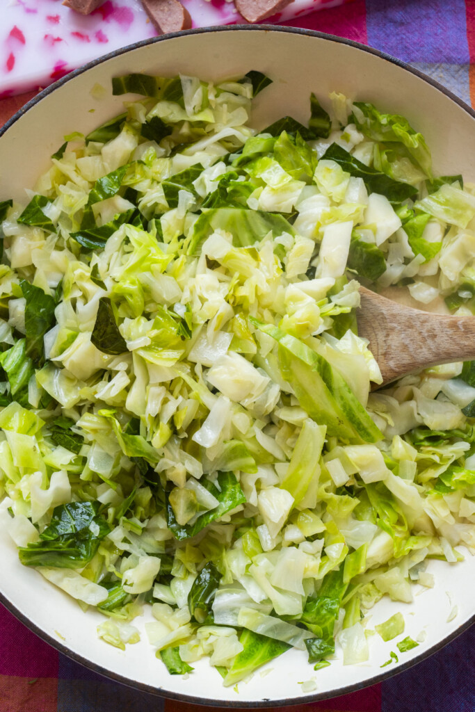 Chopped green cabbage being sautéed in a pan with a wooden spoon, showing vibrant green and light yellow hues.