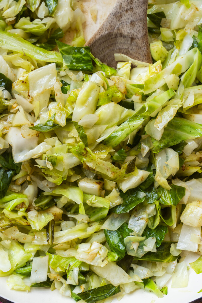 Chopped cabbage sauteed with spices in a pan, being stirred with a wooden spoon. The cabbage appears lightly cooked, with some pieces retaining bright green color.