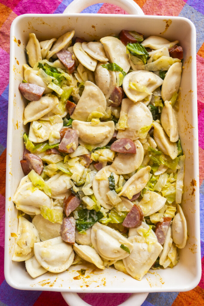 A casserole dish filled with pierogi, sliced sausage, and cooked cabbage, all mixed together, sits on a colorful, patterned tablecloth.