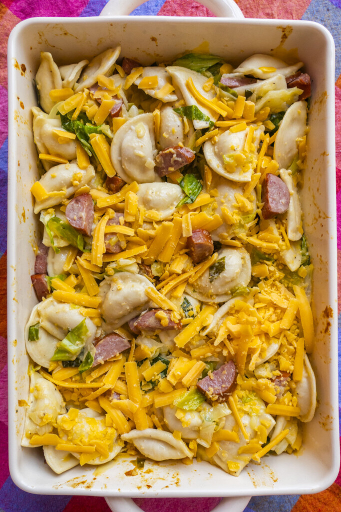 A casserole dish filled with cooked pierogies, sliced sausage, shredded cheddar cheese, and chopped green vegetables, all mixed together.