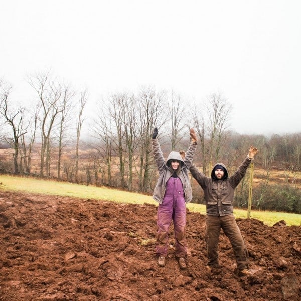 Two people in winter clothing stand on muddy ground with their arms raised, surrounded by bare trees and a foggy, overcast landscape in the background.