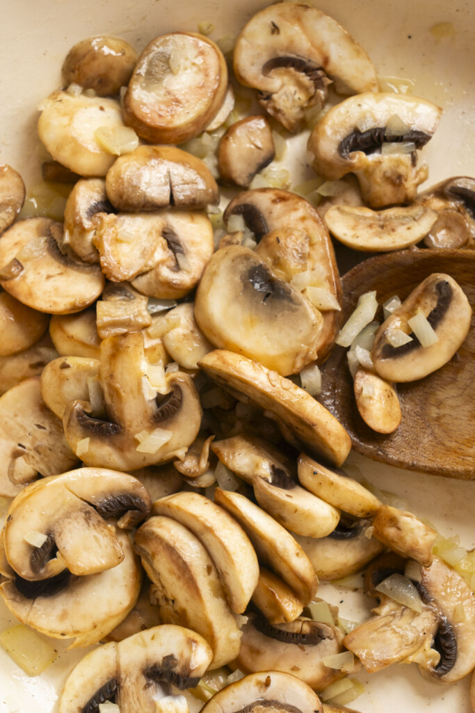 Sliced mushrooms being sautéed with chopped onions in a pan, with a wooden spoon visible among the vegetables.