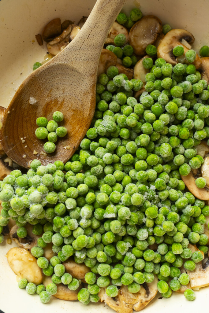 A wooden spoon rests in a pan filled with sliced mushrooms and a generous amount of frozen green peas, ready to be cooked together.