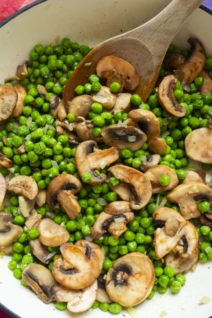 A close-up of sautéed mushrooms and buttery green peas being stirred with a wooden spoon in a white pan.