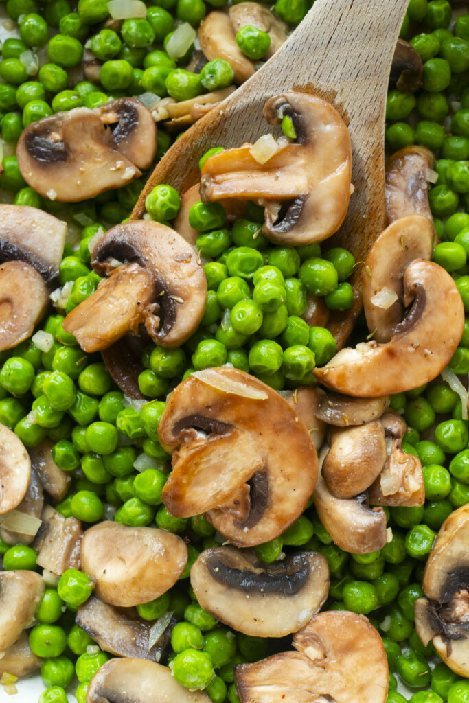 Close-up of a wooden spoon stirring cooked green peas and sliced mushrooms. The vegetables appear lightly saut&eacute;ed, with some small pieces of onion visible among the peas and mushrooms.