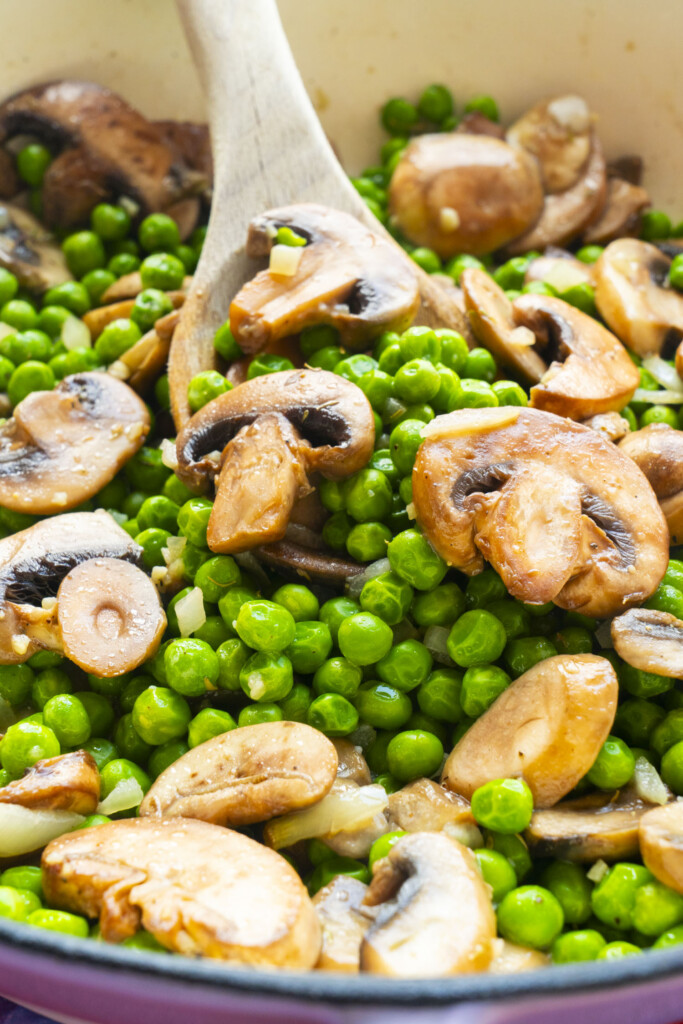 Sautéed mushrooms and green peas in a pan with a wooden spoon, showing the vegetables mixed and lightly cooked.