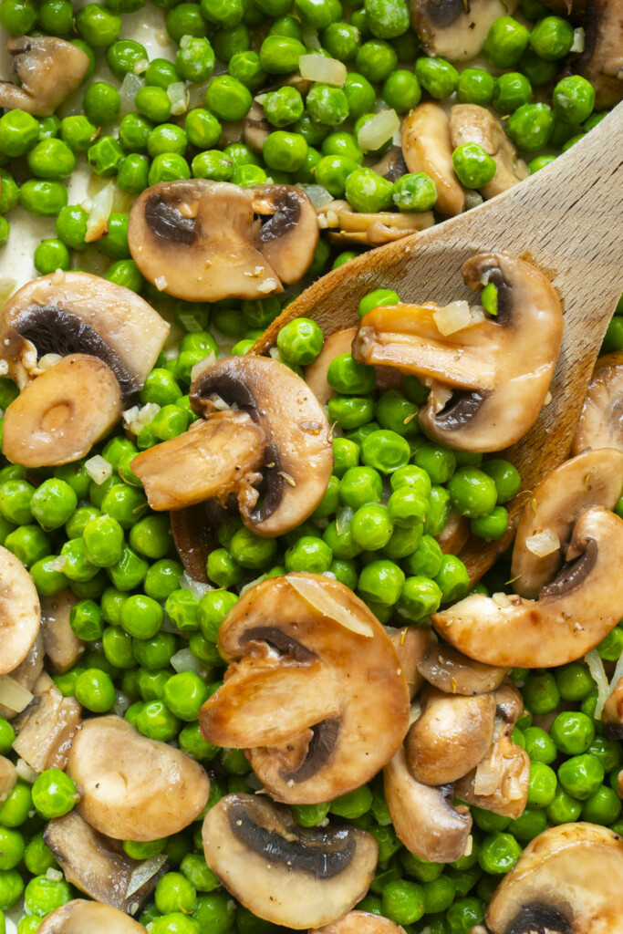 Sautéed mushrooms and green peas mixed together, with a wooden spoon partially visible stirring the vegetables.