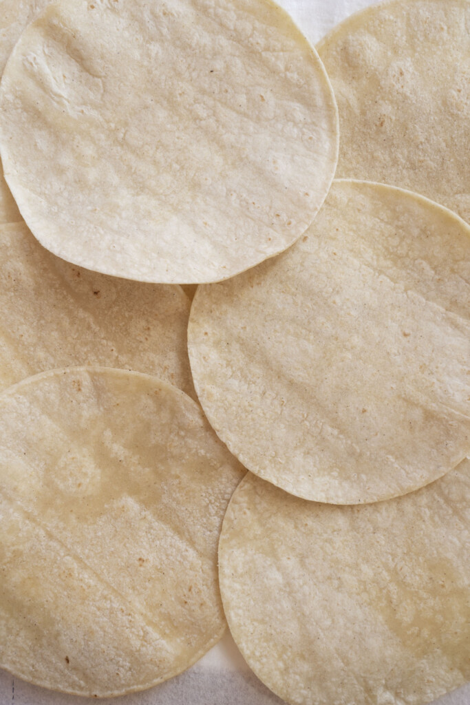 Several cooked corn tortillas overlap each other, forming a textured pattern with their round, flat shapes and pale yellow color. The tortillas have a slightly rough surface and are arranged casually.