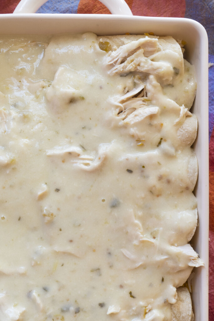 A close-up of a baking dish filled with shredded chicken and tortillas covered in a creamy white sauce, ready to be baked.