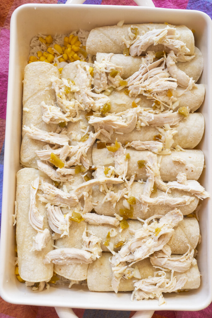 A baking dish filled with rolled tortillas topped with shredded chicken, diced green chiles, and corn, ready to be baked into an enchilada casserole.