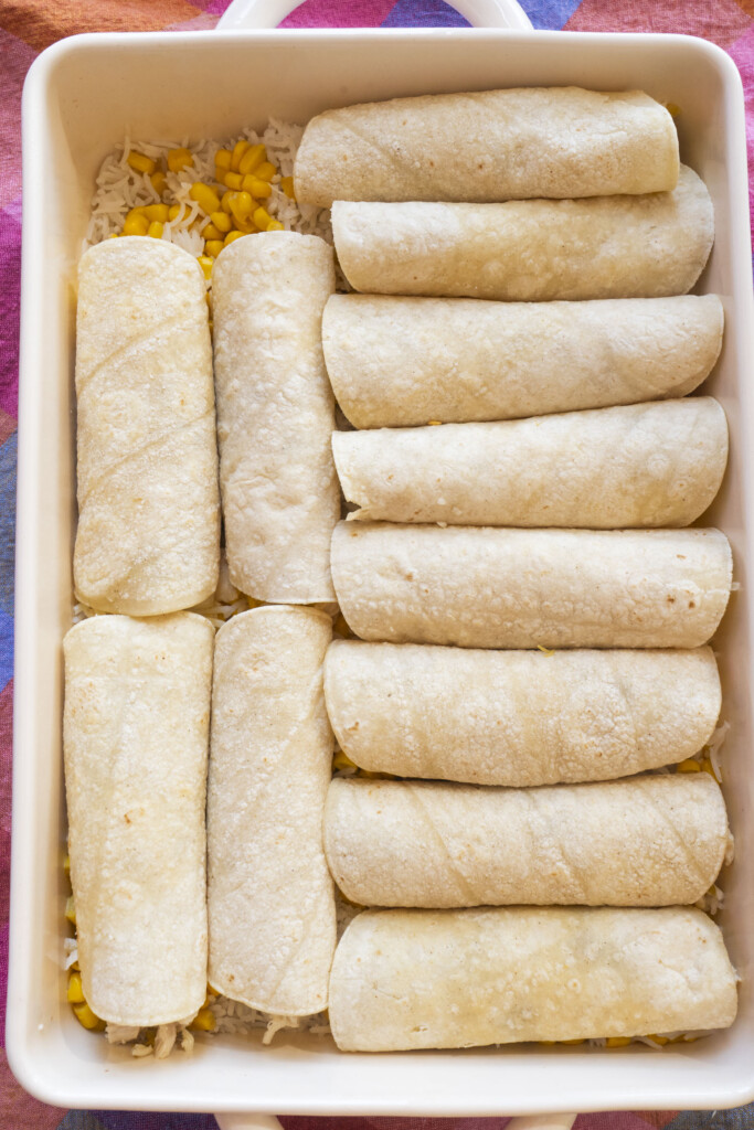 A white baking dish filled with twelve rolled tortillas neatly arranged in rows, placed on a colorful cloth. Some corn kernels and rice are visible underneath the tortillas.