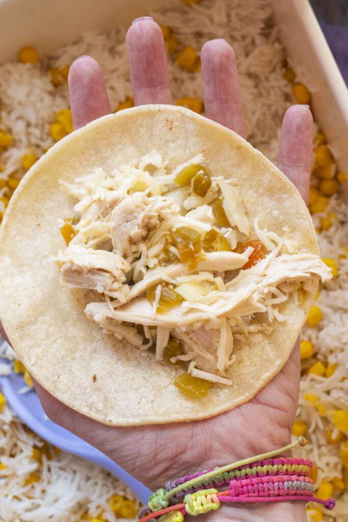 A hand holding a corn tortilla filled with shredded chicken, cheese, and diced green chilies. The background shows cooked white rice mixed with corn. The person is wearing colorful woven bracelets.