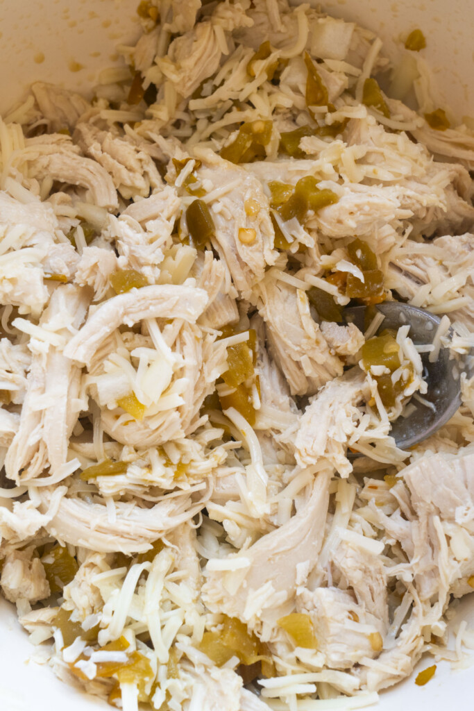 Shredded cooked chicken mixed with cheese and chopped green chilies in a bowl, with a metal spoon partially visible.