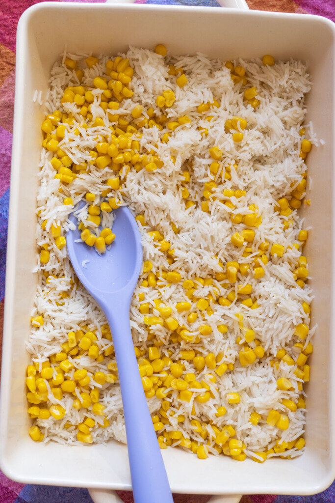 A rectangular dish filled with white rice and yellow corn kernels, with a light purple spoon resting inside. The dish is placed on a colorful, patterned surface.