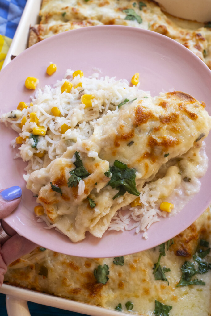 A pink plate with white rice mixed with yellow corn and baked creamy chicken topped with melted cheese and fresh herbs. A hand with blue nail polish holds the plate above a baking dish of the same chicken dish.