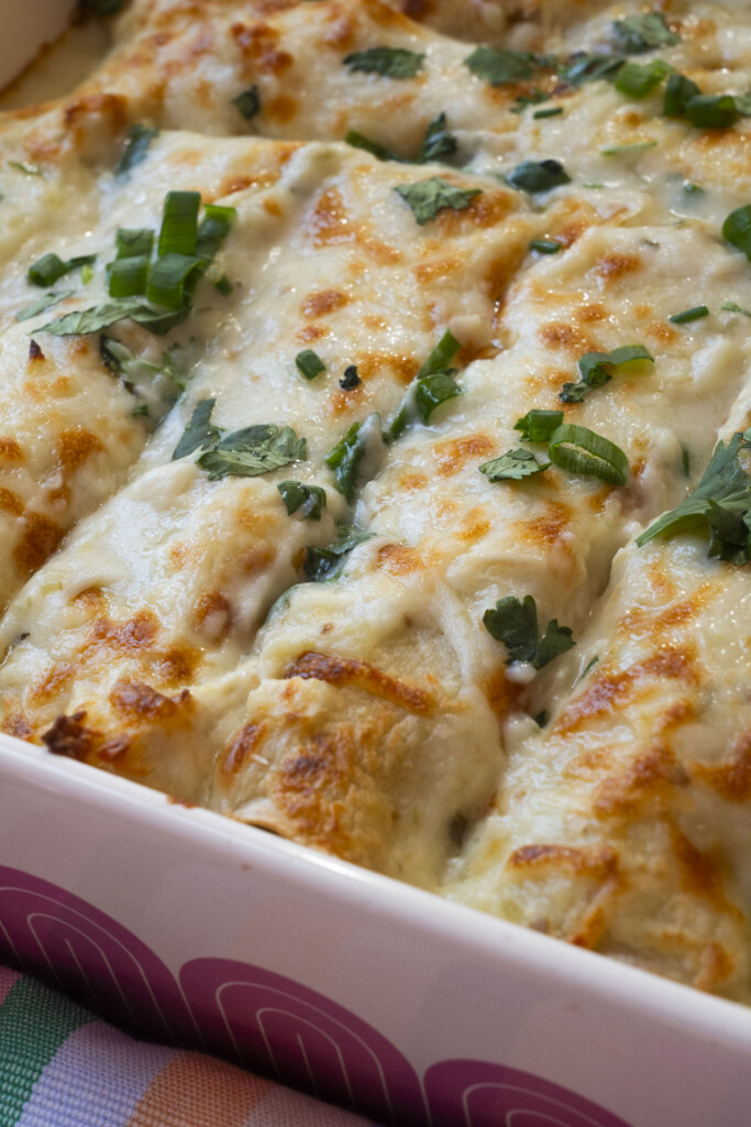 Close-up of a casserole dish filled with cheesy, baked enchiladas topped with melted cheese and garnished with chopped fresh herbs.